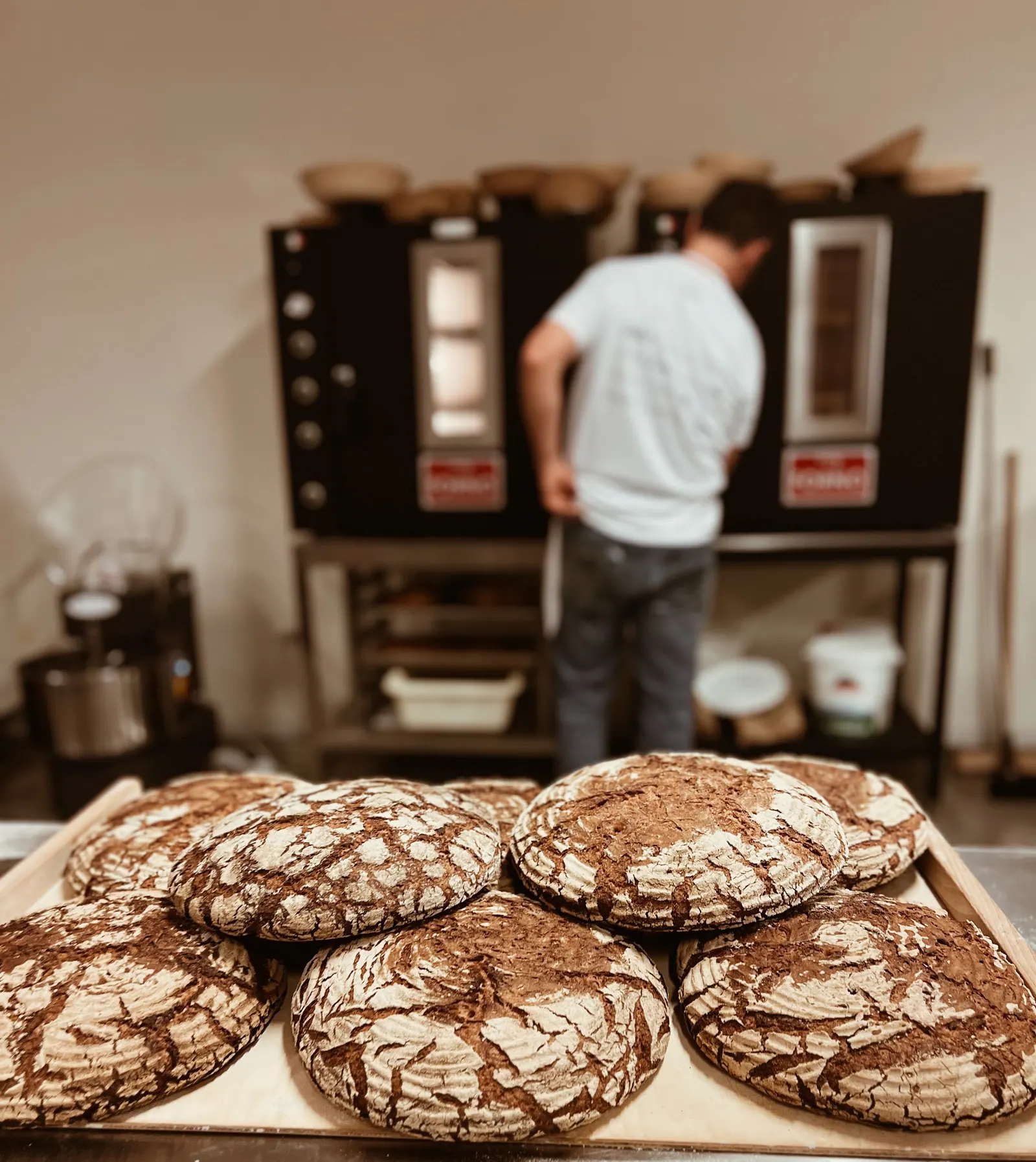 Large dark rye loaves on a board, a baker at two MyForno ovens behind