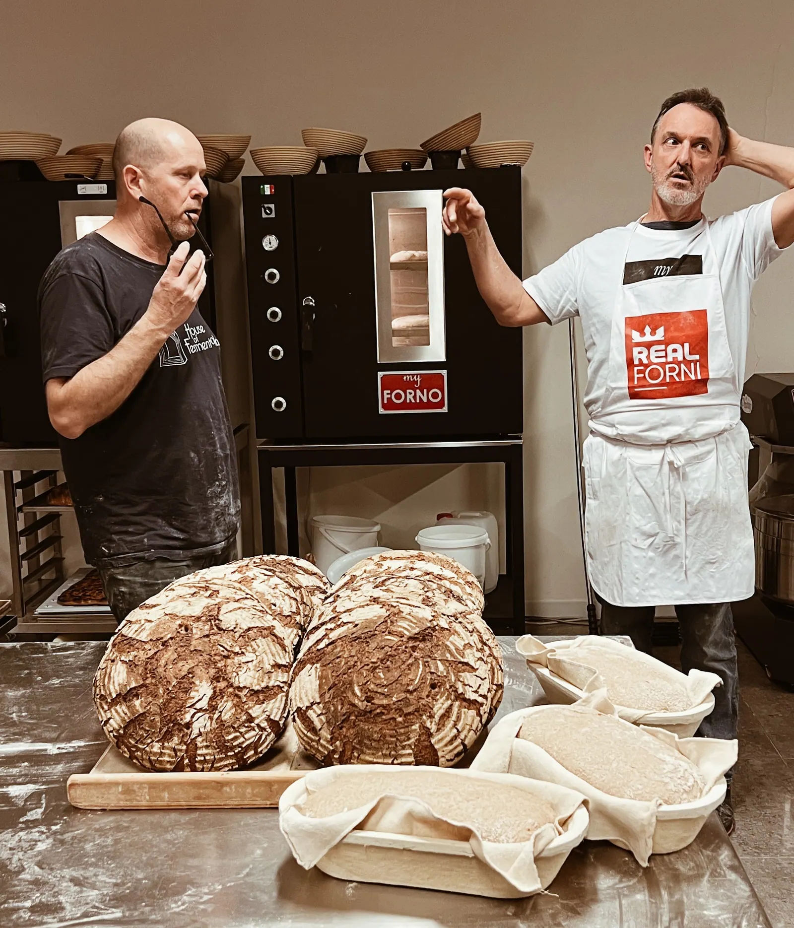Two bakers by two MyForno ovens, dark rye loaves on the bench in front