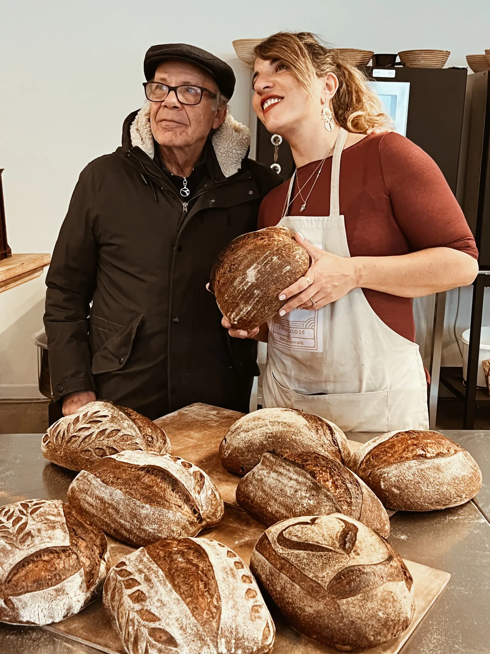 An older baker and a baker in an apron holding a loaf behind a board of their bread.