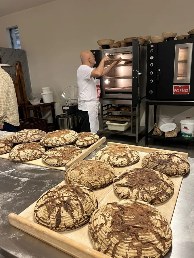 A board of scored rye loaves, a baker at a black oven behind