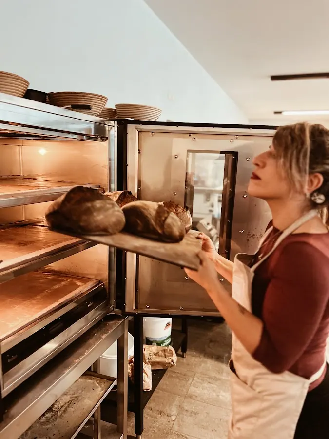 A baker sliding a peel of dark sourdough loaves into a deck oven
