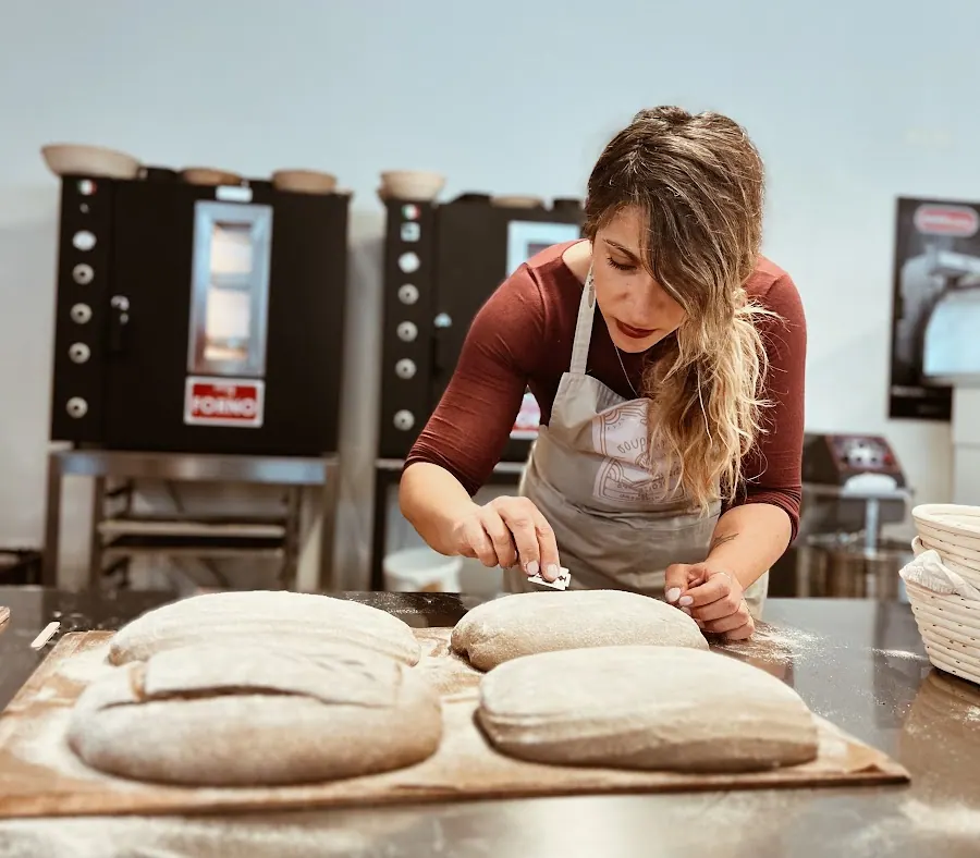 A baker scoring three loaves on trays, two black MyForno ovens behind